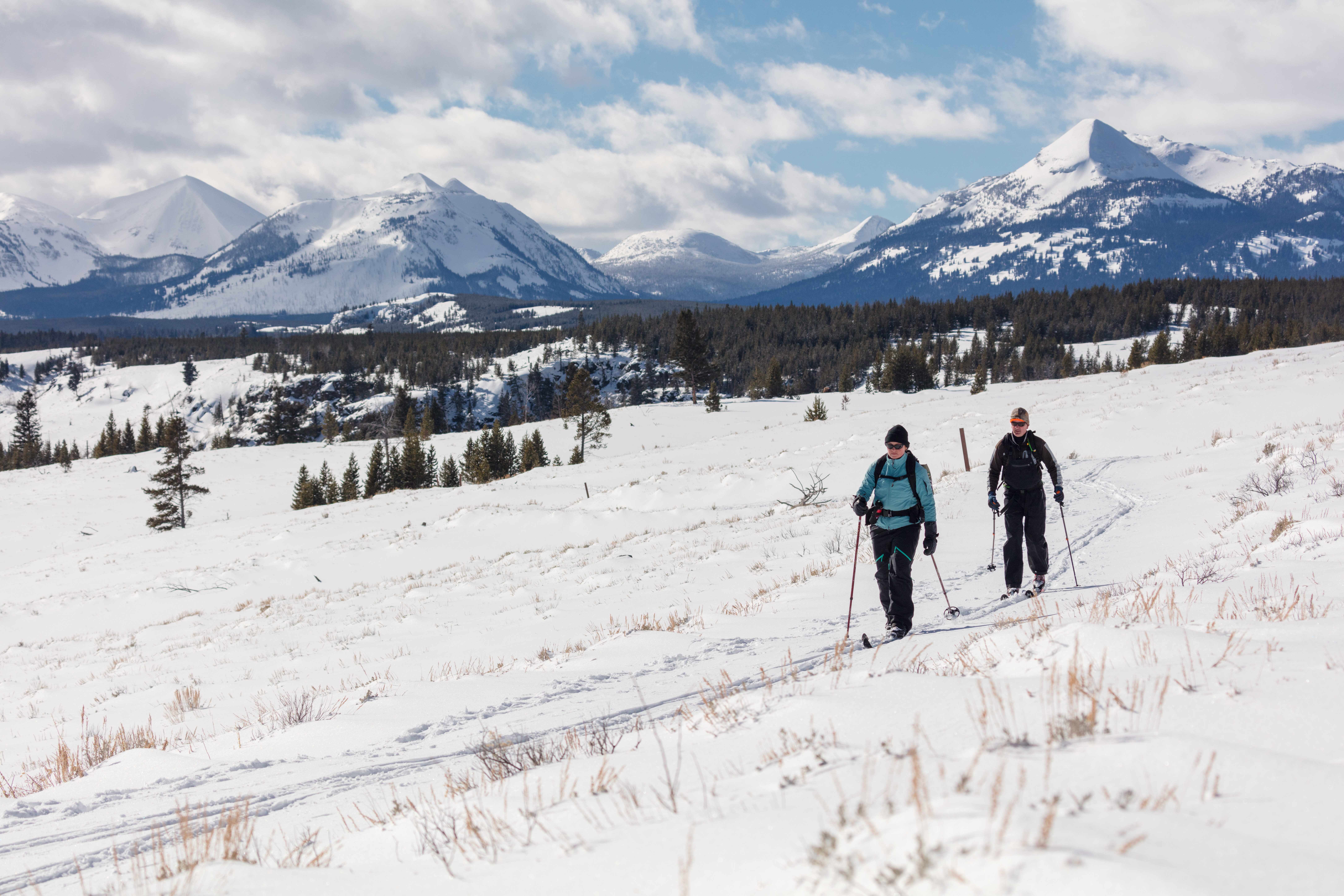 Two skiers travel across a flat meadow with mountains in the distance.
