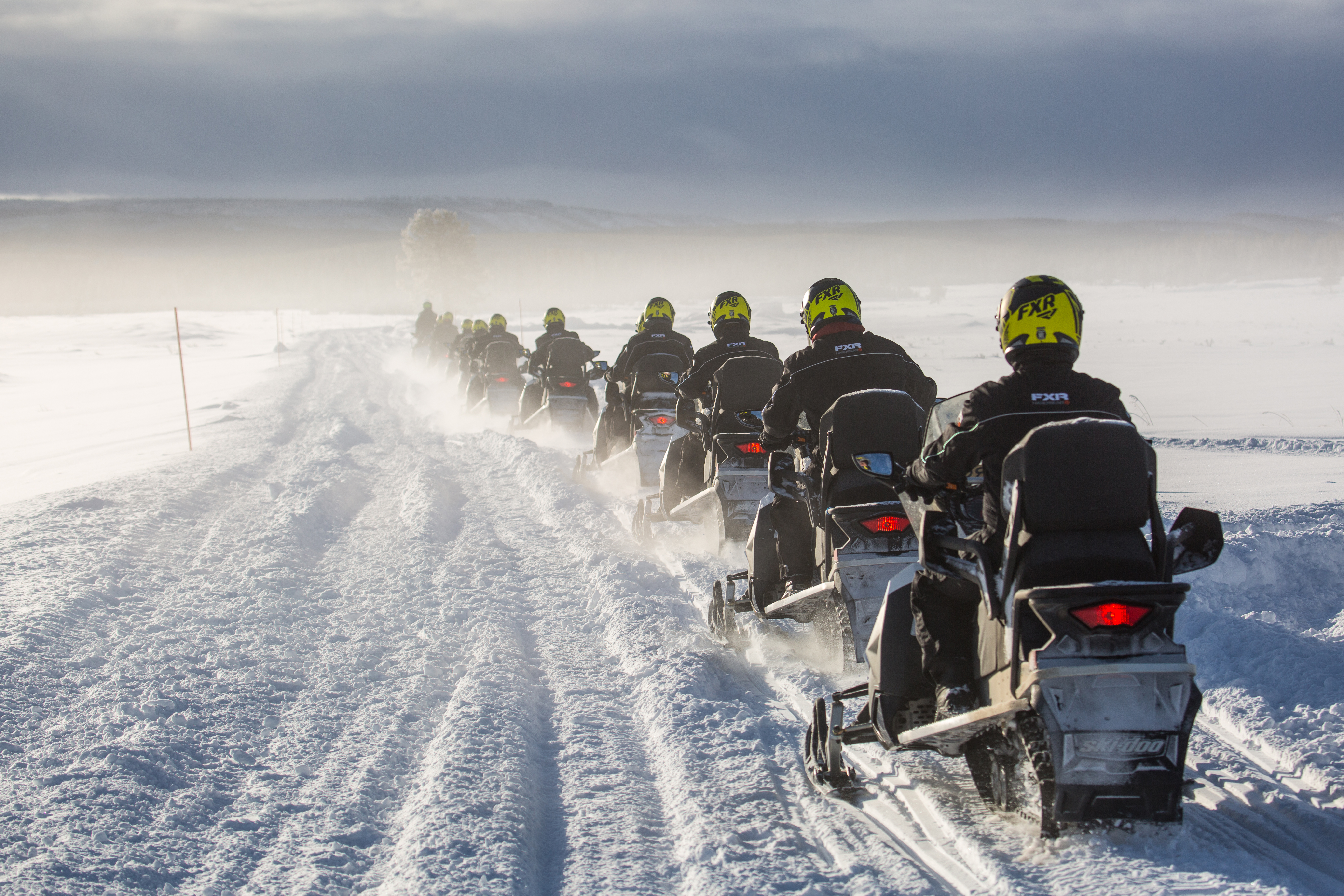 A line of snowmobiles move along a snowy track that leads across a snow-covered valley.