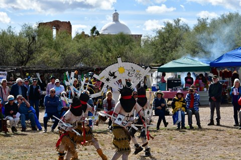 apache crown dancers with large head pieces dance with audience watching and church in background