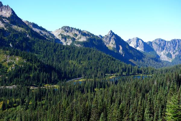 A road curves through a forested valley next to a lake and under a mountain range.