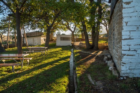 The setting sun casts light and shadows over buildings and a fenced yard with trees.