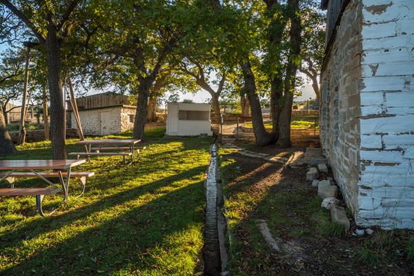 The setting sun casts light and shadows over buildings and a fenced yard with trees.
