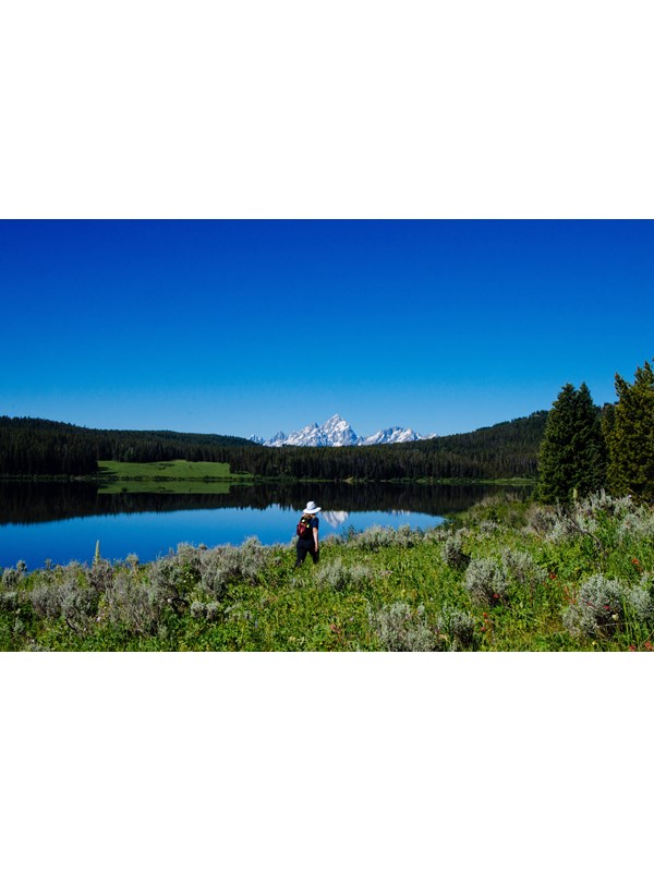 A hiker walks along the lakeshore with mountains in the background.