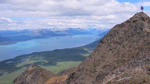 A woman stands on top of a rocky mountain looking out towards the turquoise water of Lake Clark