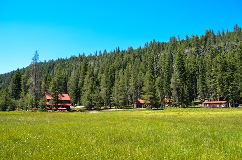 A two-story red and brown lodge and three cabins at the base of a hillside fronted by a meadow