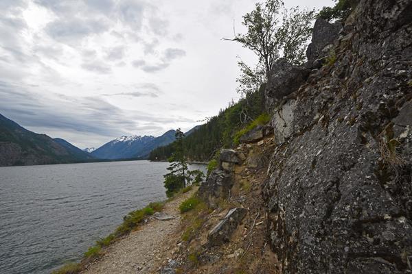 A dirt path follows the shore of a large lake, with snow-capped peaks in the distance.
