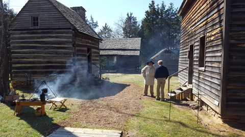 Living history interpreter stands at historic farm with an outdoor fire.