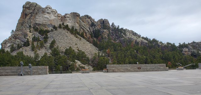 The four carved presidents on Mount Rushmore above a pine-covered talus slope, seen from the wide granite paving of the Grand View Terrace