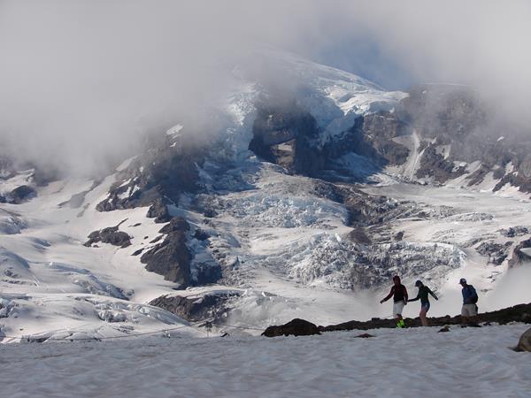 Three hikers crossing a large patch of snow with Mount Rainier in the background.
