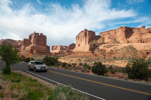 A tan car drives through Arches, sandstone features are visible in the back.