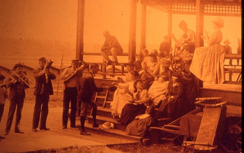 A group of people in period clothing listening to a band play with the lake in the background.