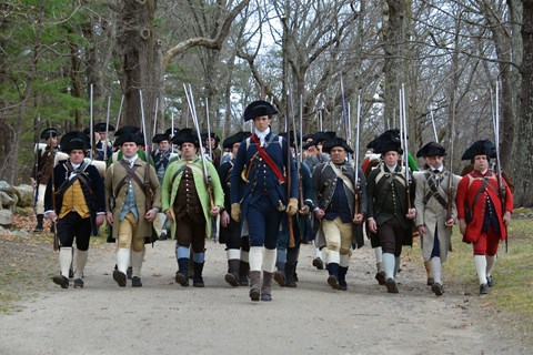 A group of Minute Men in 1770's clothing marching down a dirt road with muskets on their shoulders