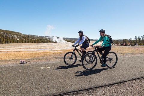 Two people bike on a paved trail past a geyser in a thermal basin.