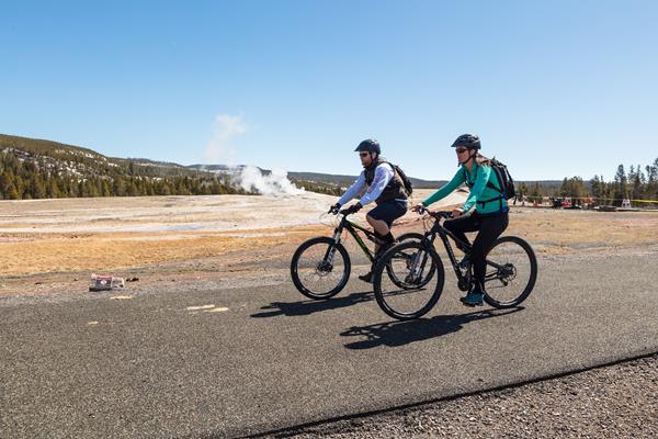 Two people bike on a paved trail past a geyser in a thermal basin.