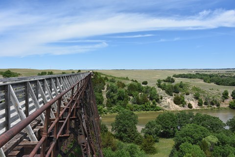 A metal bridge with wood walkway extends across a green landscape with a river below.
