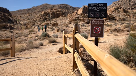 A dirt trail between split rail fences with three hikers in the distance heading towards mountains.