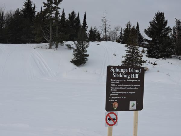 Brown sign titled 'Sphunge Island Sledding Hill' in front of a hill covered with snow.