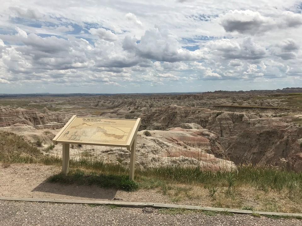 A wayside exhibit stands at the edge of drop off into badlands formations