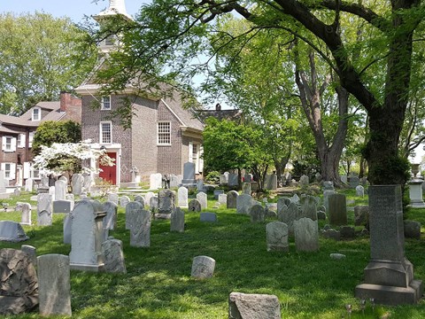 Headstones are scattered across a pastoral churchyard. The brick church sits behind them.