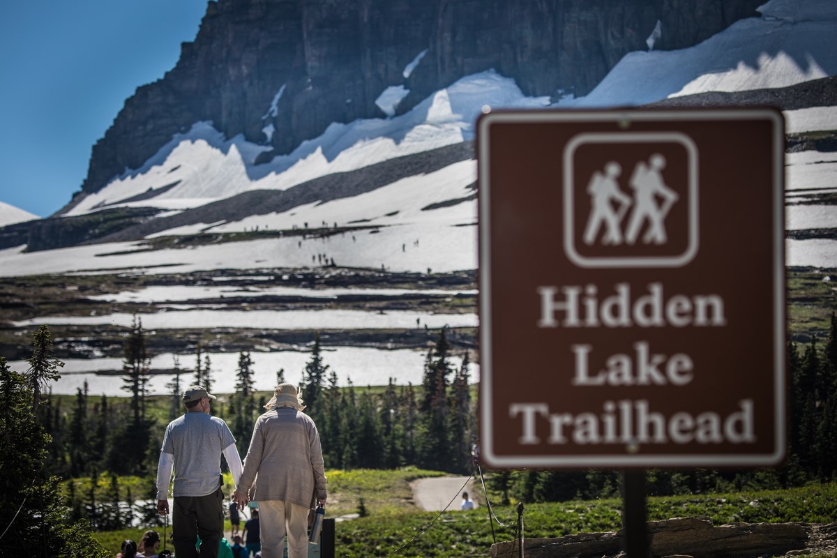 A sign that reads "Hidden Lake Trailhead" in front of hikers and a mountain.