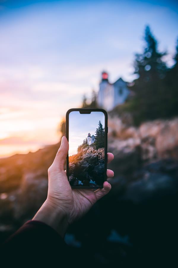 A hand holds a phone poised to snap a photo of a lighthouse bathed in sunset on a rocky coastline.