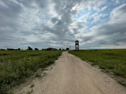 A gravel road through a grassy field towards distant buildings.