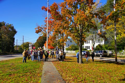 A ranger in NPS uniform stands under a flagpole surrounded by fall foliage speaking with visitors