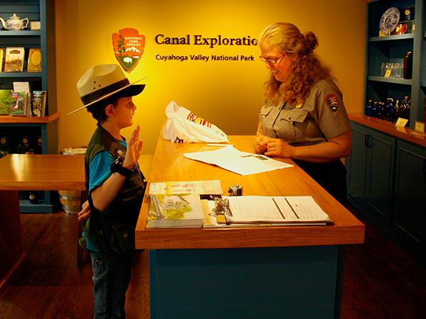 A child wearing a ranger hat holds up their right hand, across a desk from a uniformed ranger.