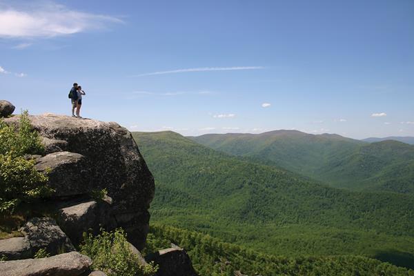 Two hikers standing on a huge boulder looking over a view of mountain ranges and a green valley