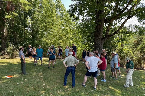 A ranger and several visitors gather on a grassy knoll surrounded by woods.