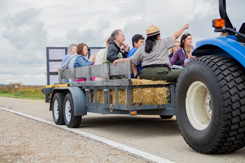 A woman in a flat hat and grey shirt points to an area over people's heads on a hay ride