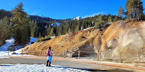 A woman on showshoes takes a photo of a large mudpot in a mountain landscape.