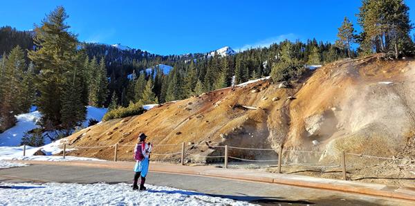A woman on showshoes takes a photo of a large mudpot in a mountain landscape.