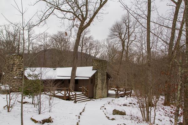 An historic cabin with a large porch sits in the middle of a snow-covered forest.
