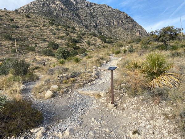 A trail sign marks the junction of three trails in a desert mountain landscape