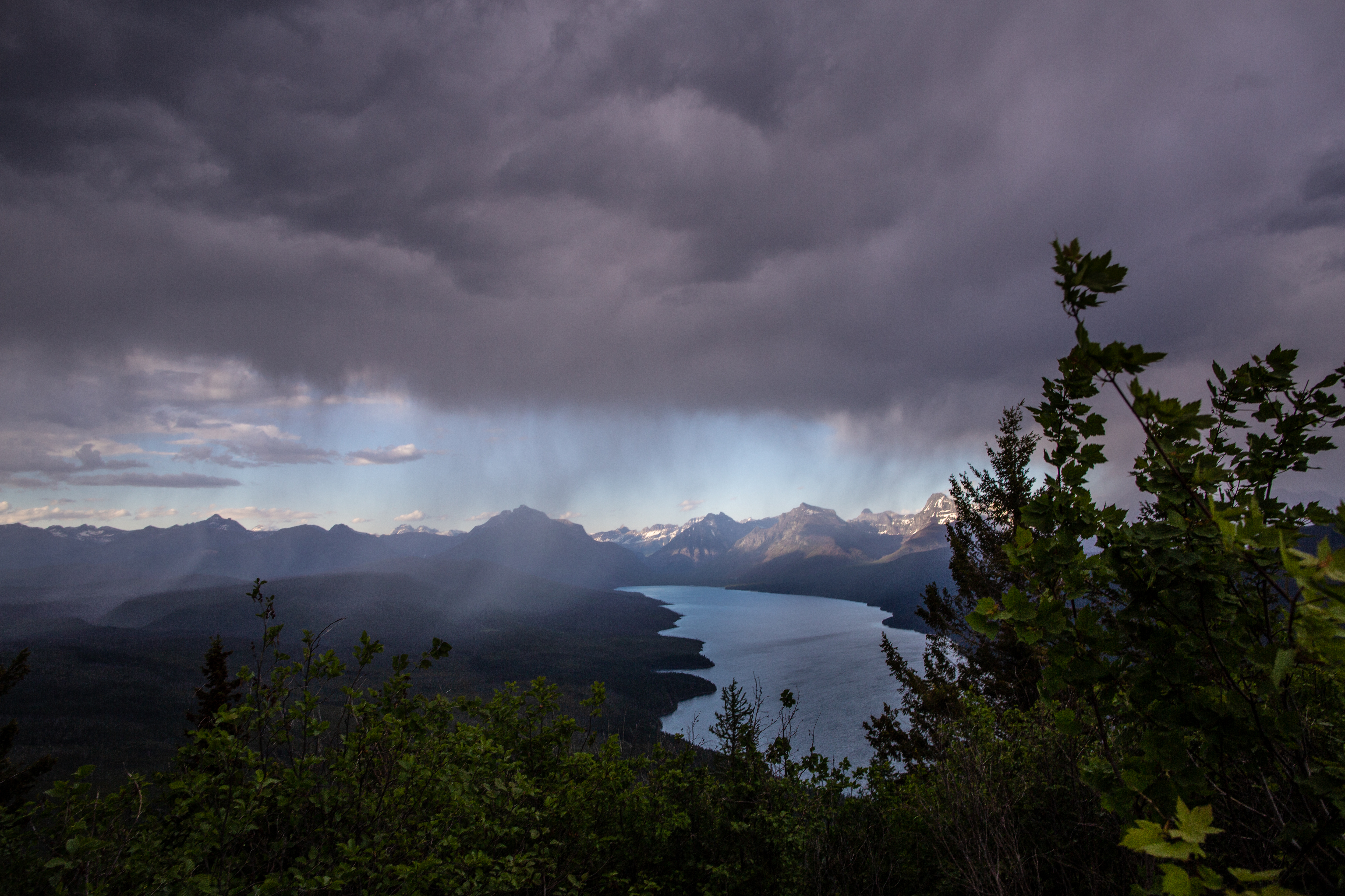 Trees in the foreground, mountains clouds, and a lake in the background.