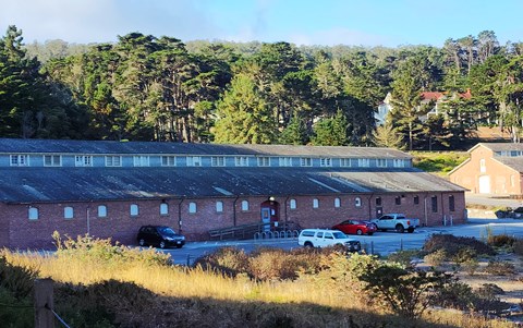 Long brick building with a parking lot in front surrounded by trees.