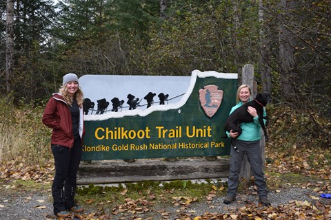 two women and a dog pose with a Chilkoot Trail sign