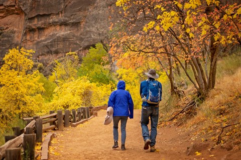 Two hikers in blue jackets walk along a sandy trail lined with a wooden rail and fall foliage.
