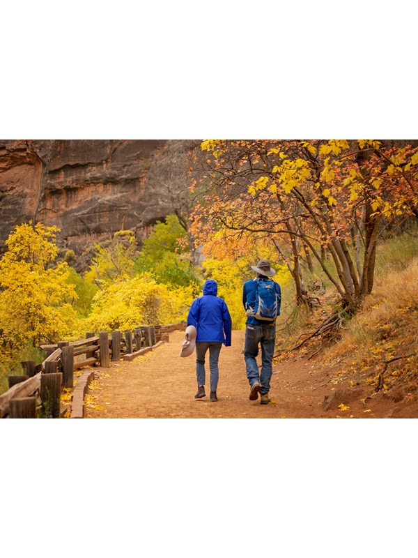 Two hikers in blue jackets walk along a sandy trail lined with a wooden rail and fall foliage.
