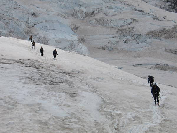 A string of climbers, darkly silhouetted against a white, glacier-covered slope, follow a boot track
