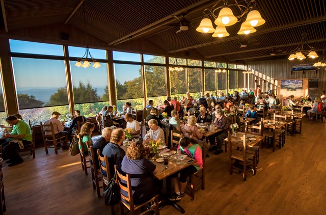 A group of diners sit at several tables in a dining room with wooden floors and large windows.