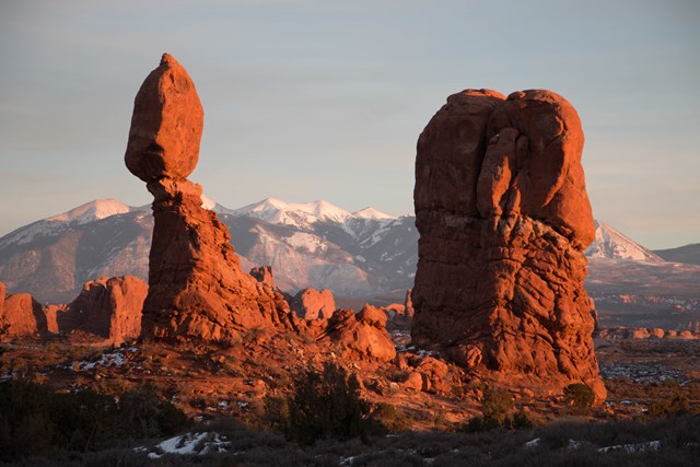 Sandstone boulder atop its pedestal glowing orange at sunset with snowy peaks behind