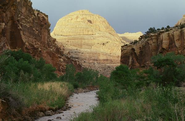 Large rounded white sandstone dome centered above a river, with green grass, red cliffs, & blue sky