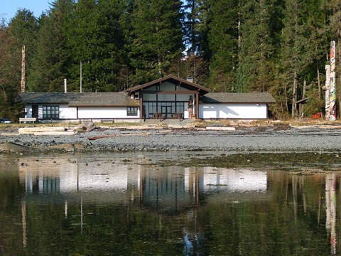 Calm water with a low, white building flanked by totem poles in the background