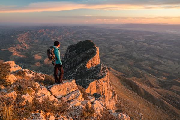 A hiker stands on a peak with lower mountains in the background at sunset