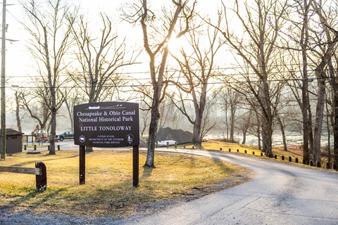 Entrance sign for the C&O Canal National Historical Park at Little Tonoloway beside a curving paved road and the river