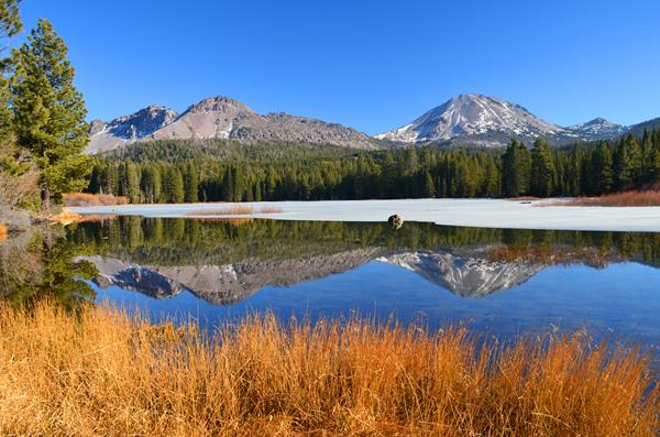 Two volcanic peaks reflected in a partially frozen lake lined by golden grass and evergreen trees.