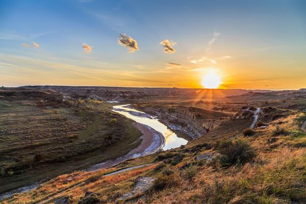 A bend in the river and a trail above it are seen at sunset. Buttes and prairies line the river.