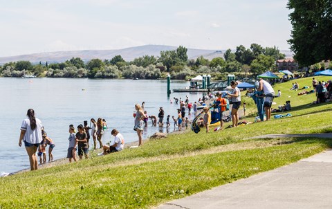 Color photograph of a river's shore with numerous people gathered on a green lawn beside it.